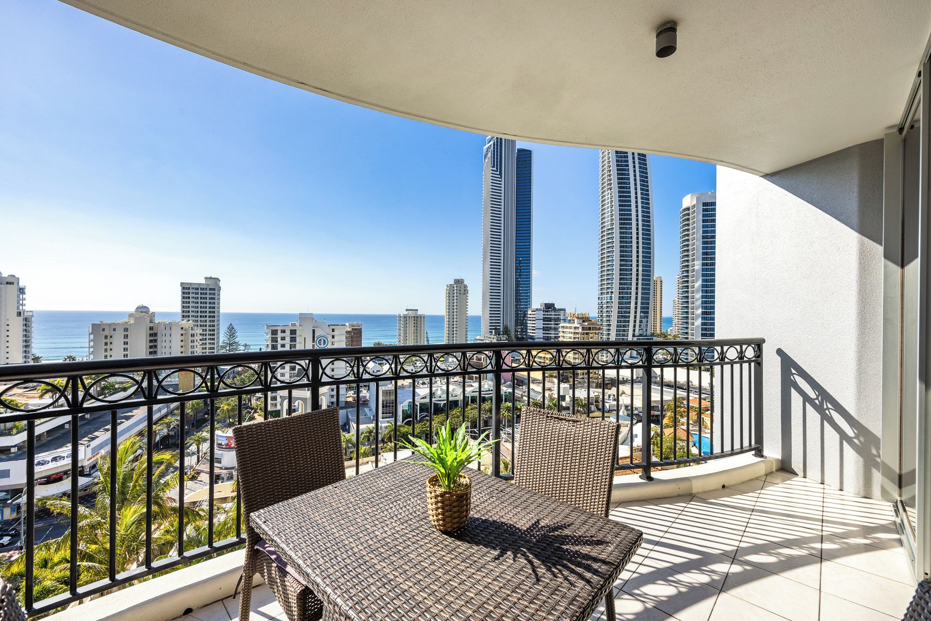 A balcony with a table and chairs, overlooking a cityscape with tall buildings and a clear sky.