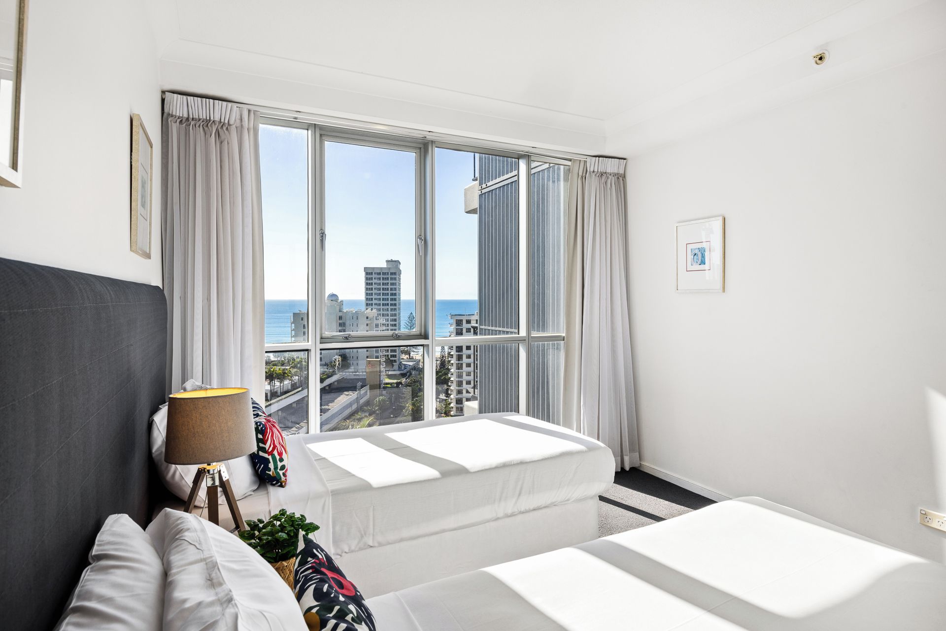 A hotel room with a large window, two beds, a bedside table with a lamp, and a framed picture on the wall.