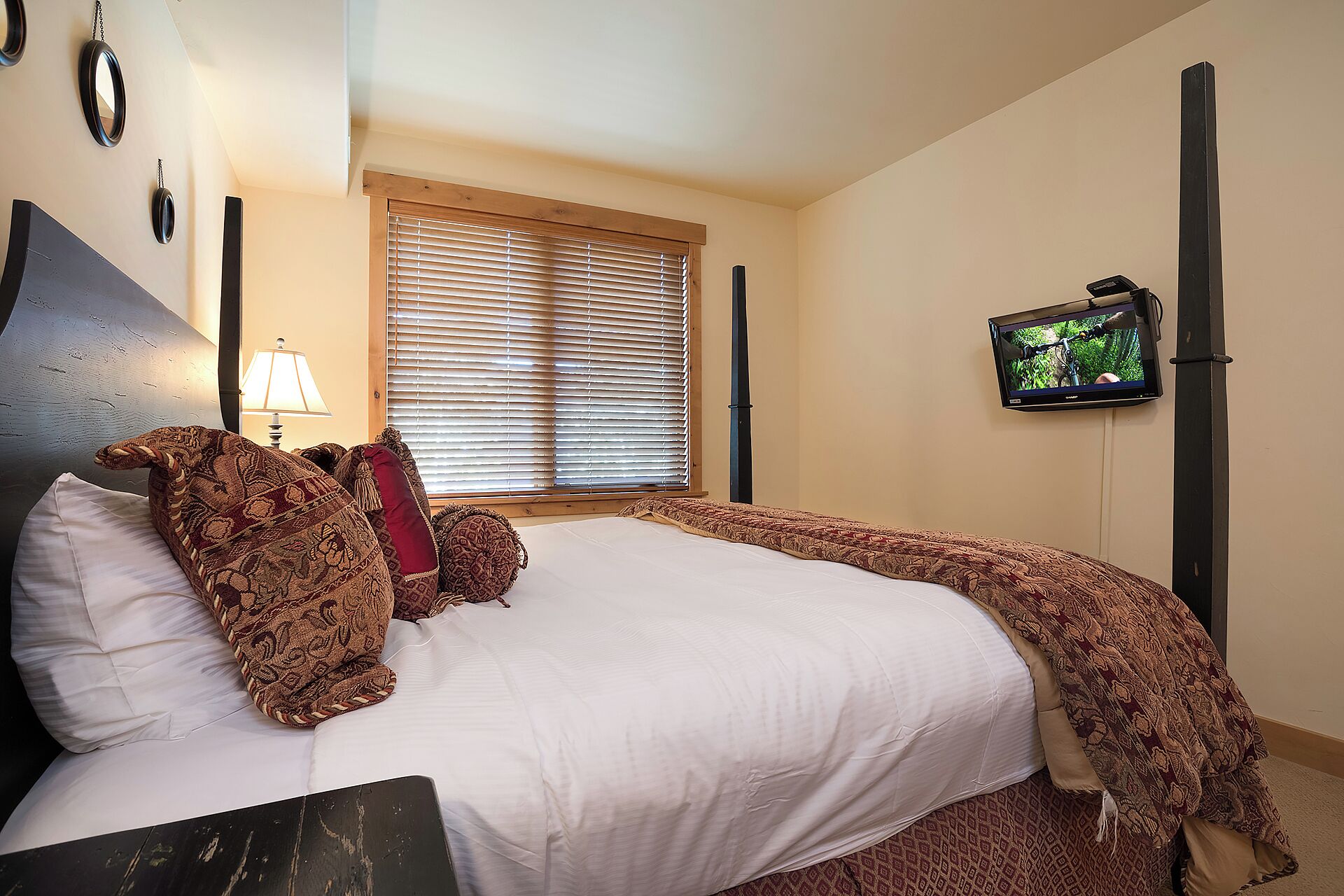 A bedroom with a bed, a television mounted on the wall, and a window with blinds.