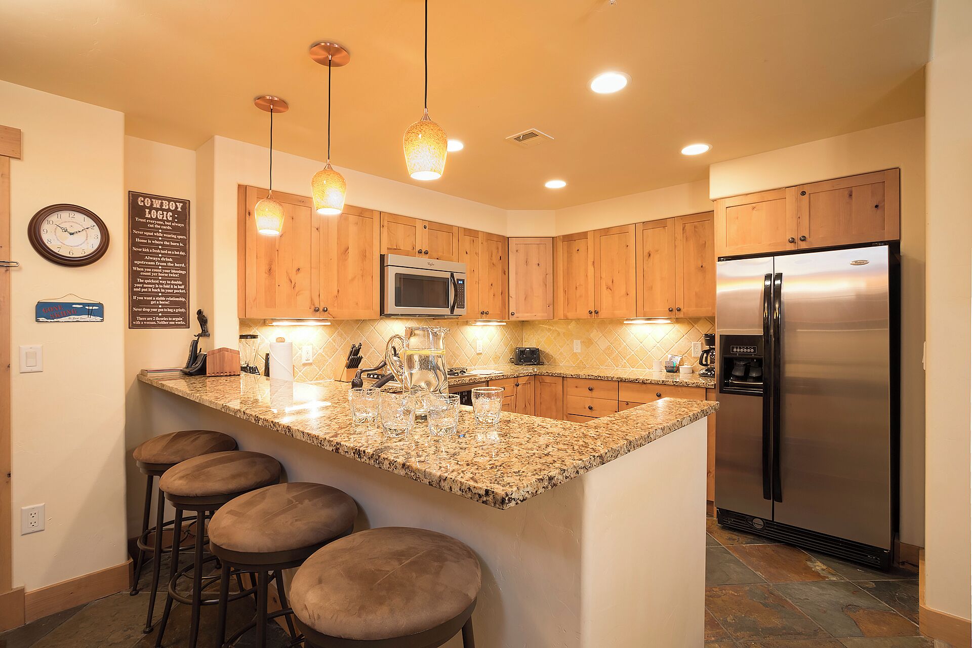 A kitchen with wooden cabinets, a stainless steel refrigerator, a granite countertop, and pendant lighting.