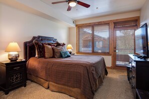 Um quarto com uma cama grande, duas mesinhas de cabeceira, um ventilador de teto e uma televisão sobre um suporte de madeira.