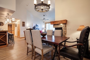 Uma sala de jantar com mesa de madeira, cadeiras estampadas, um candelabro e uma lareira de pedra.