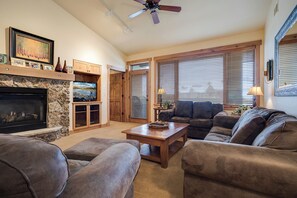 A living room with a fireplace, a television, a ceiling fan, and large windows with blinds. - AL4304 | Top Floor with Vaulted Ceilings | Views | 4 Pools | Air Chilling (Steamboat Springs)