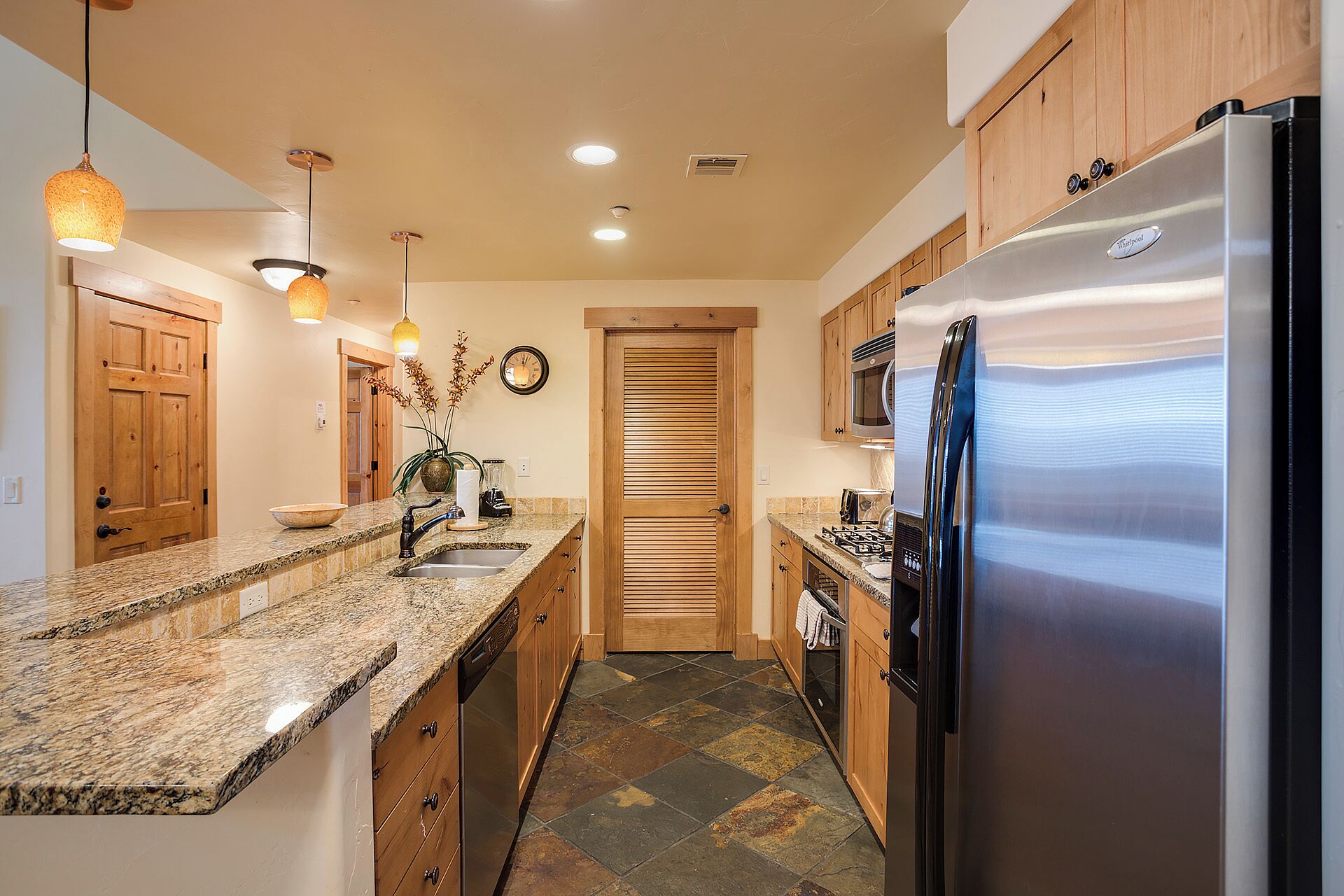 A kitchen with granite countertops, wooden cabinets, a stainless steel refrigerator, and a stone-tiled floor.