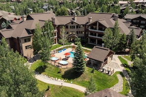 Aerial view captures the property nestled in a tree-lined residential community, featuring a central pool and landscaped grounds surrounded by mountain lodge-style architecture. - AL4305 | Top Floor Residence | Beautiful View | Air Chilling | 4 Pools (Steamboat Springs)