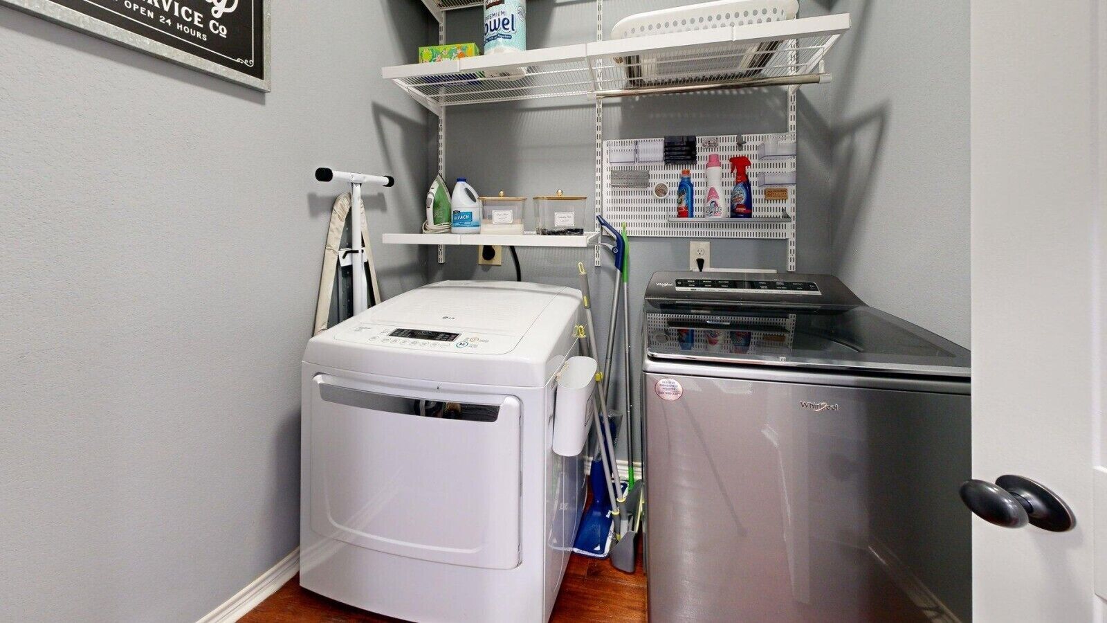 Un cuarto de lavandería con lavadora y secadora de carga frontal, repisas con productos de limpieza y un letrero que indica servicio las 24 horas.