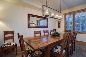 A dining area with a wooden table, six chairs, a chandelier, a mirror, and a window with blinds. - BL6102 | Unforgettable Mtn Getaway | 4 Pools | Walk to Mountain | Air Chilling (Steamboat Springs)