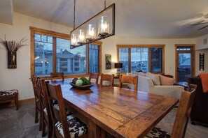 Un comedor espacioso con una mesa y sillas de madera, una araña y ventanas amplias con persianas.
