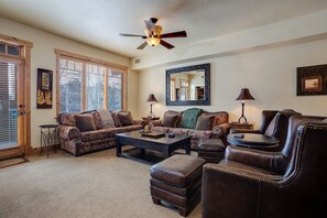 A living room with a sofa, ottoman, coffee table, and a leather armchair. There is a ceiling fan, a mirror, and a lamp on a side table. - BL6106 | Mountain Residence | Private Patio with Hot Tub | Air Chilling (Steamboat Springs)