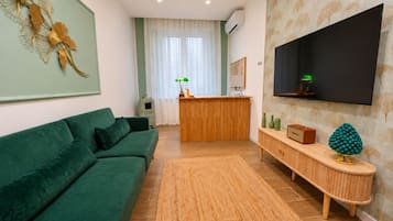 A living room with a green sofa, a wooden reception desk, a flat-screen TV mounted on the wall, and a decorative plant on a console table.