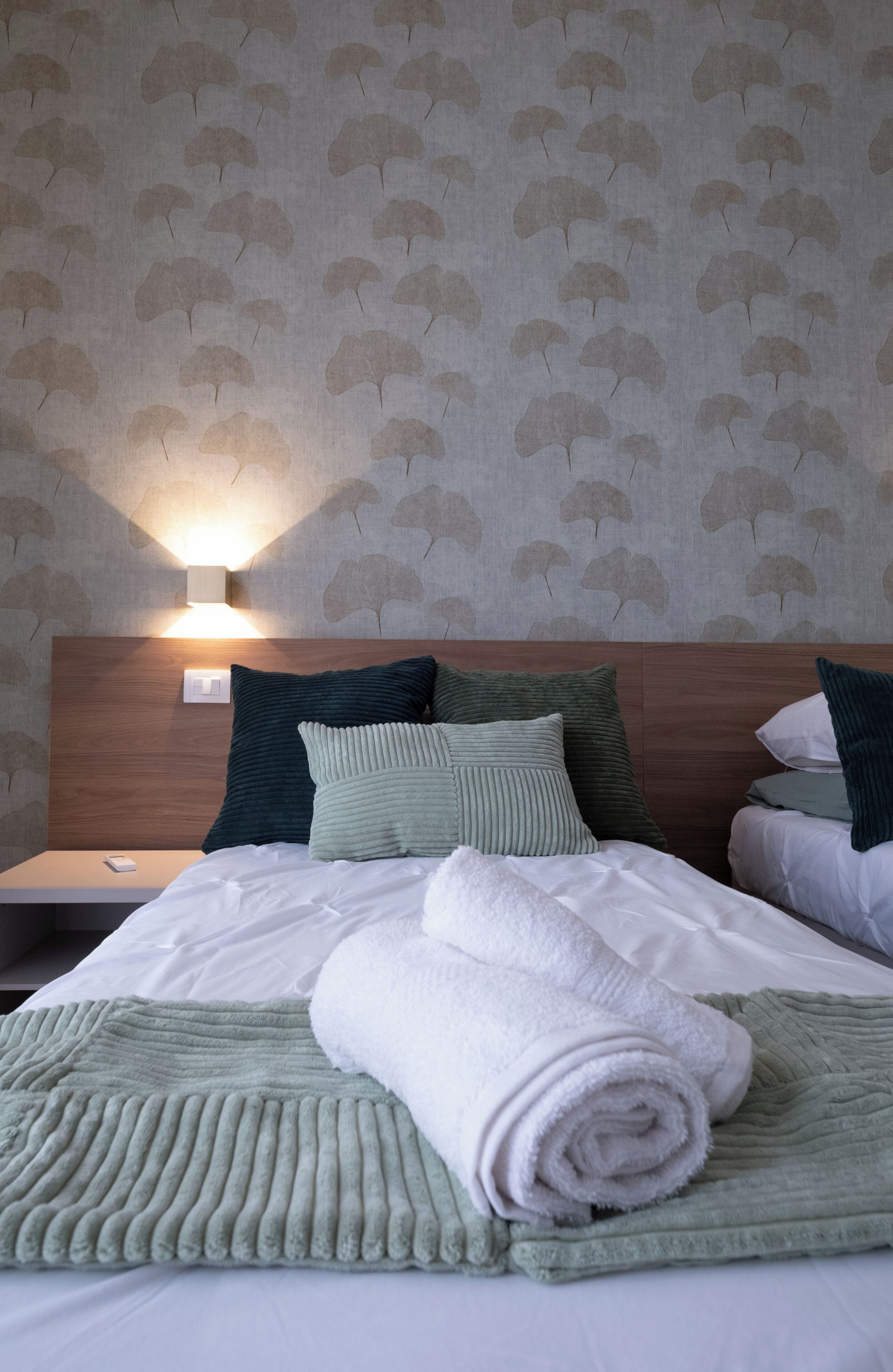 A neatly made bed with a folded towel and a green blanket, a wooden headboard, and a wall with a leaf pattern.