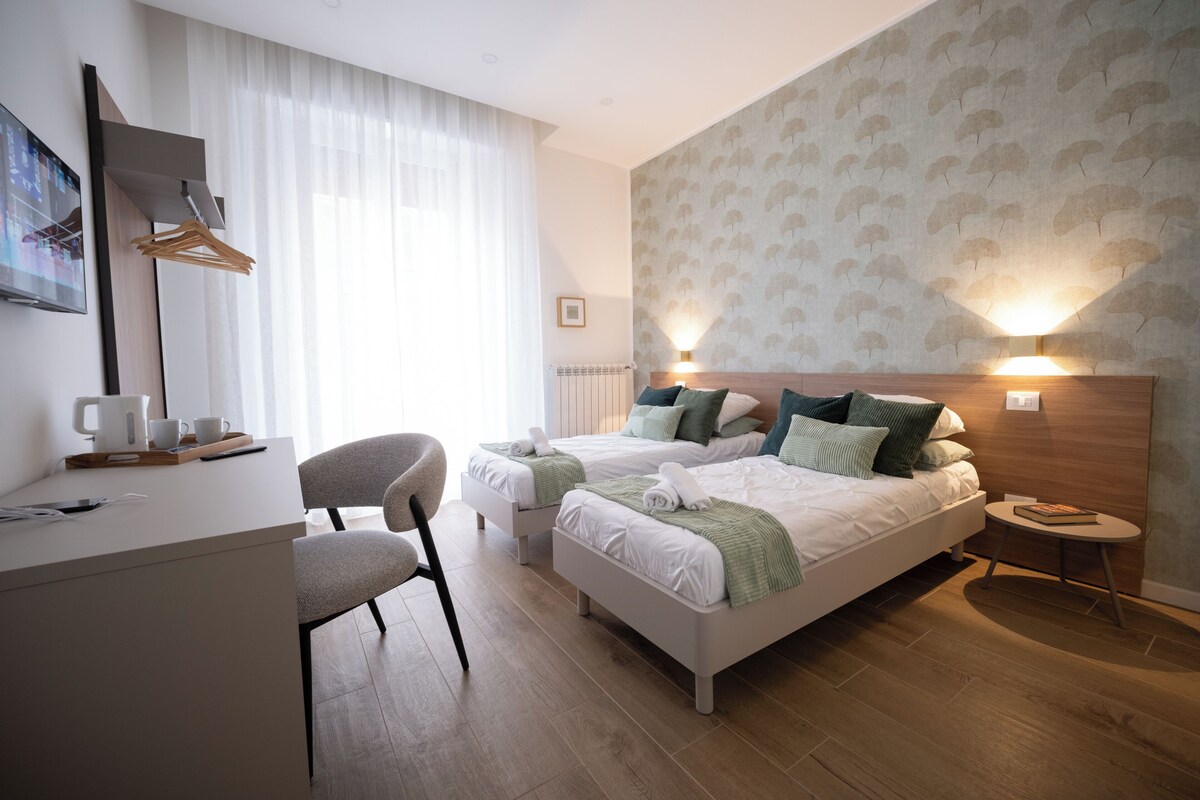 Ett hotellrum med två sängar, ett skrivbord, en stol, ett litet bord och en vägglampa.
