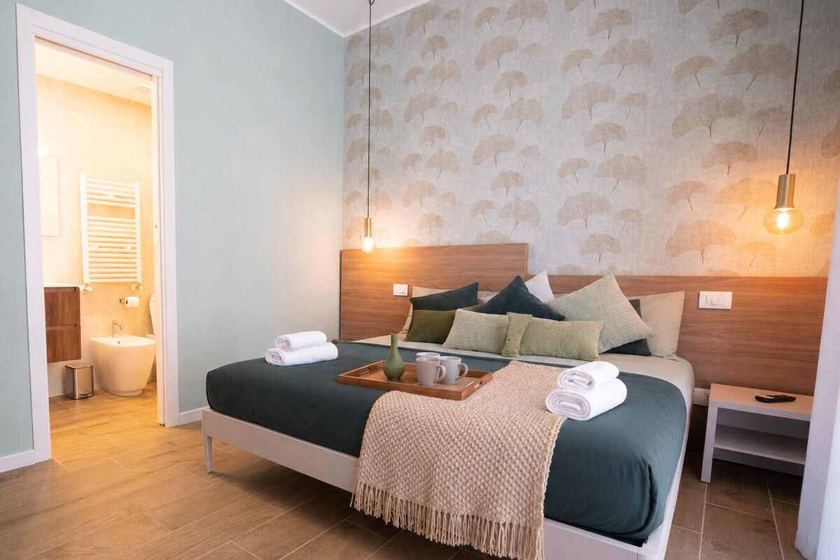 Ett hotellrum med en säng, sängbord, ett litet bord, en väggmonterad lampa och en dörr som leder till badrummet.