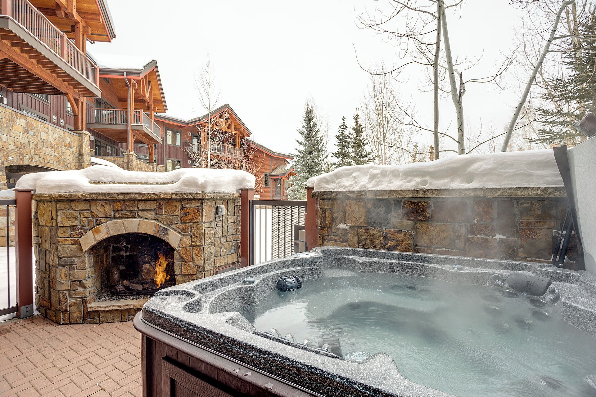 Une cheminée en pierre avec un feu de bois, un jacuzzi entouré de pierres, et un bâtiment en bois en arrière-plan.