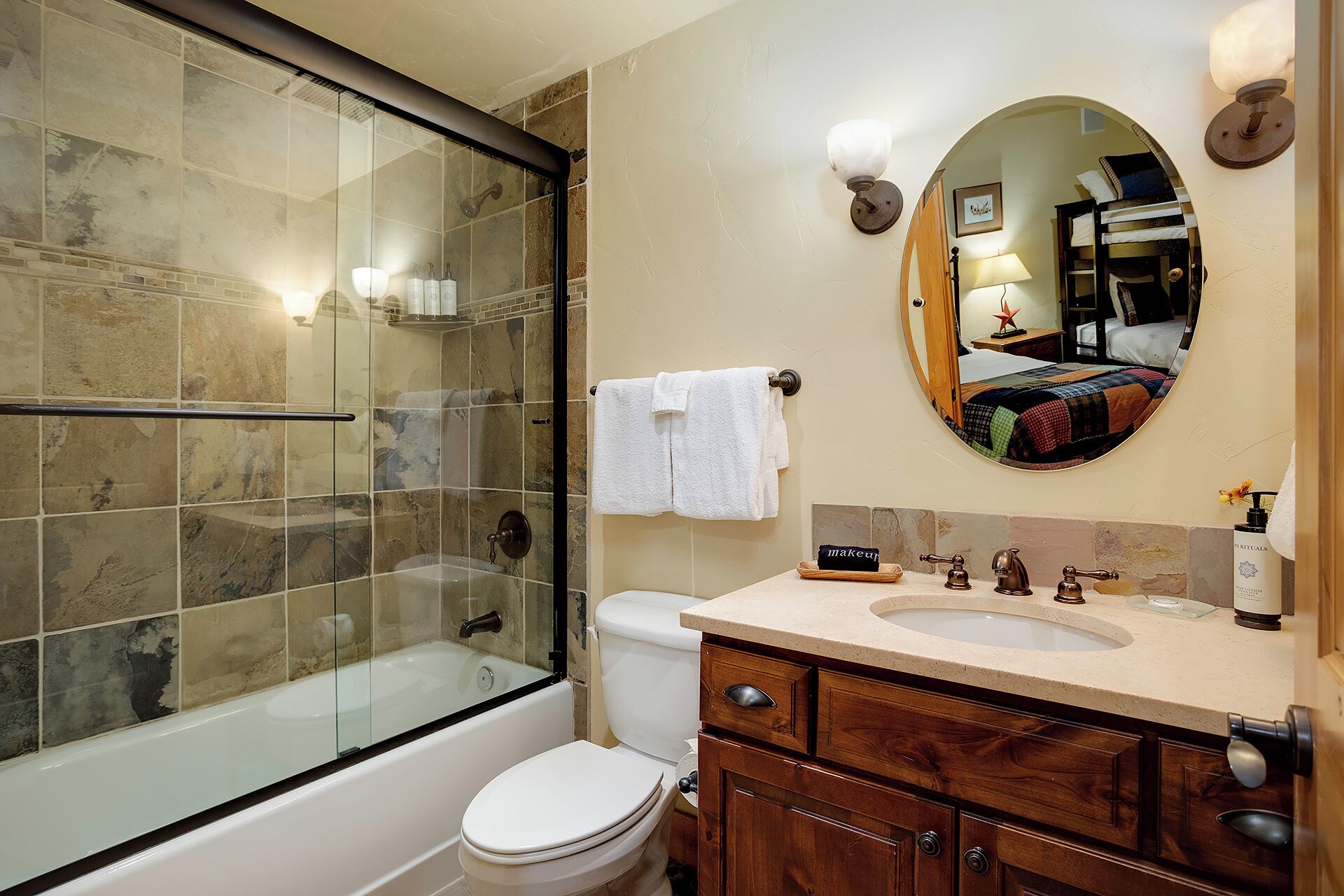 Un bagno con doccia vetrata, un water, un lavandino con mobile contenitore e uno specchio che riflette una camera da letto.
