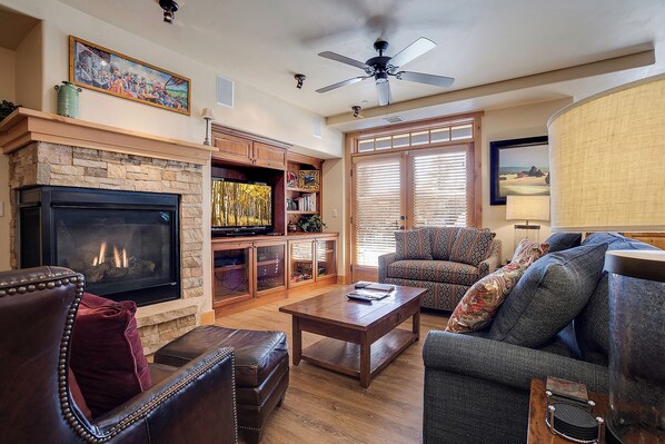 A living room with a fireplace, a television, a ceiling fan, and a large window with blinds. - BL6111 | Private Courtyard with Hot Tub | Air Chilling | Pools | Wood Flooring (Steamboat Springs)