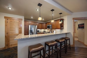 A kitchen with wooden cabinets, a stainless steel refrigerator, a microwave, and a granite countertop. There are three stools and a hanging light fixture. - BL6112 | Cozy Mtn Condo | Private Courtyard and Hot Tub | Air Chilling (Steamboat Springs)
