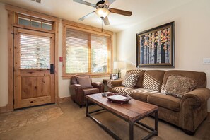Un salon avec une porte en bois, deux fenêtres équipées de stores, un ventilateur de plafond, un tableau encadré, un canapé marron, une table basse en bois et un fauteuil en cuir marron.