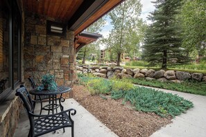 A stone building with a patio area featuring a table and chairs, surrounded by a landscaped garden with a stone wall and trees. - BL6116 | Charming Garden Apartment | Walk to Mtn | Air Chilling | 4 Pools (Steamboat Springs)