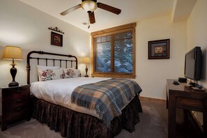 Ein Schlafzimmer mit Bett, Deckenventilator, Fenster mit Jalousien, Fernseher und Schreibtisch.