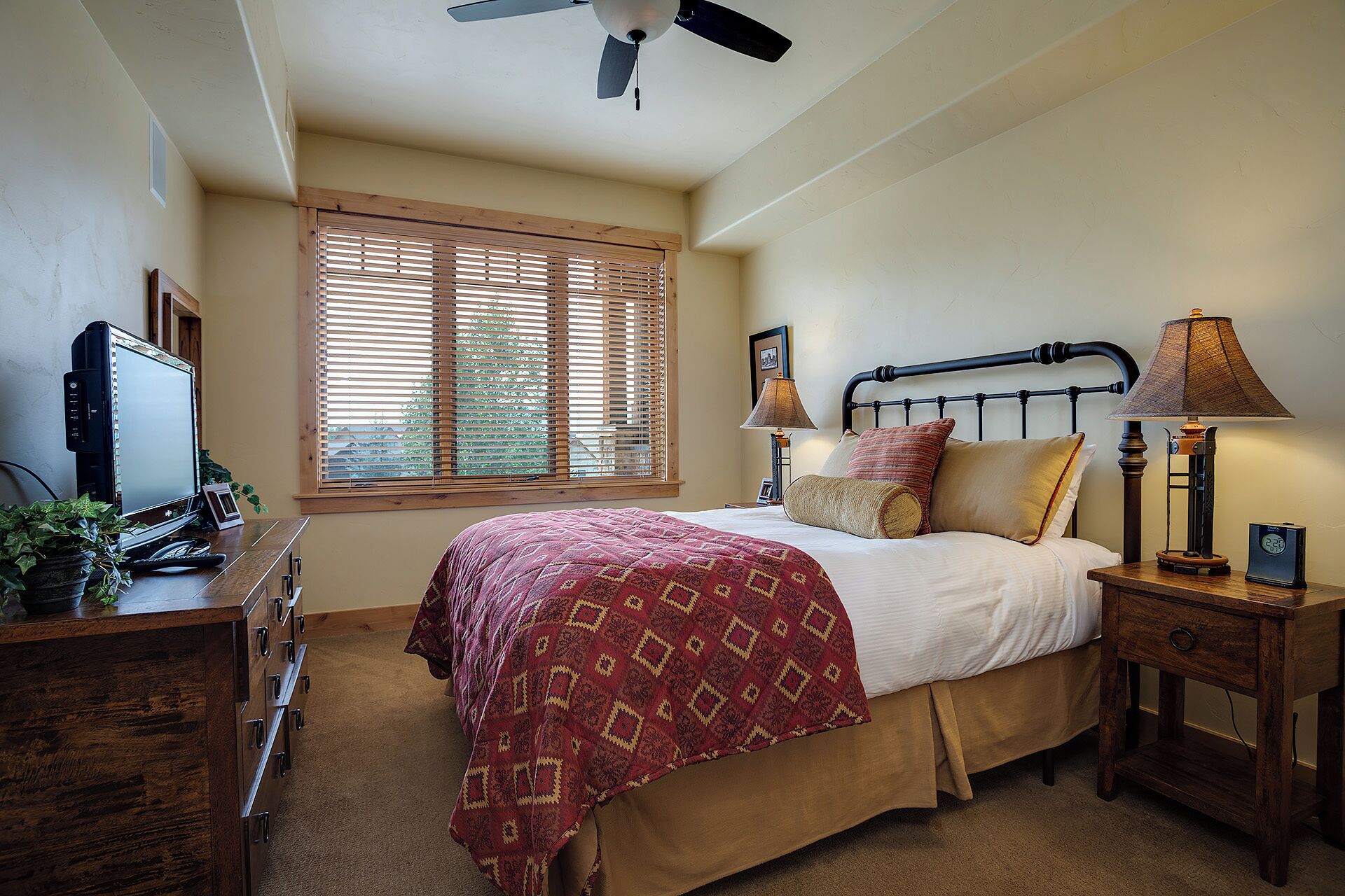 A bedroom with a bed, bedside lamps, a desk with a computer, and a window with blinds.