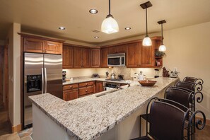 A modern kitchen with granite countertops, stainless steel appliances, wooden cabinets, and bar seating. - BL6203 | Beautiful Sunset Views| 10 Hot Tubs | Air Chilling | 4 Pools (Steamboat Springs)
