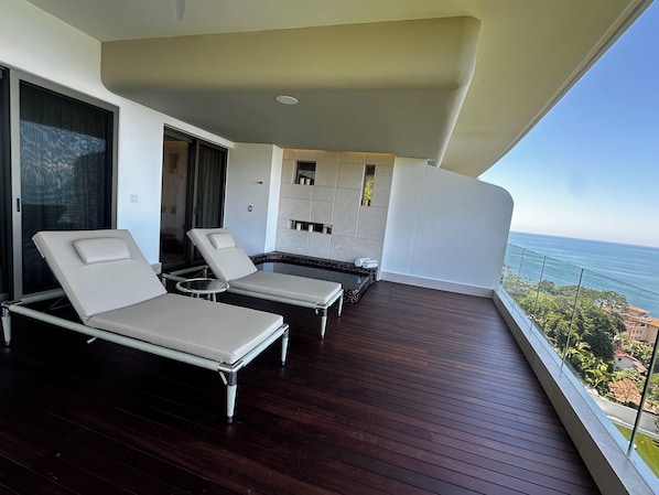 A balcony with two sun loungers and a small table, overlooking a body of water and a coastal landscape.