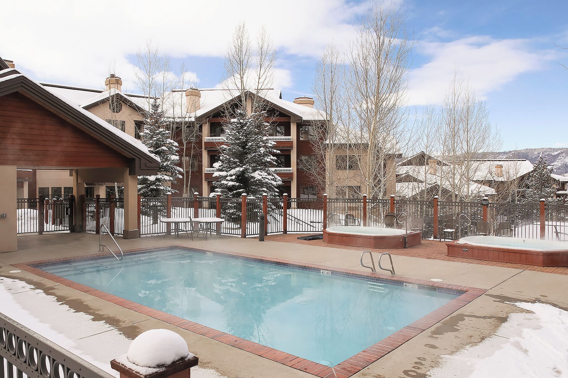 A pool area with a hot tub, surrounded by a wooden deck and snow-covered trees.
