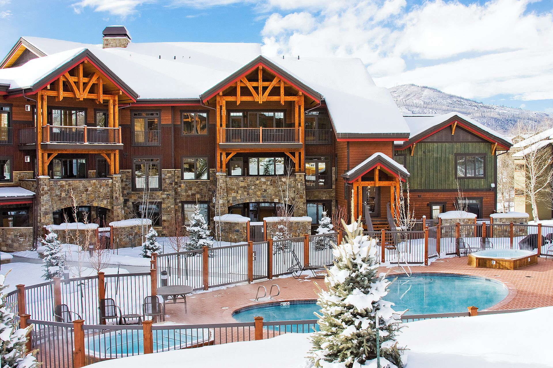 A multi-story lodge with a pool area, surrounded by snow-covered trees and mountains.
