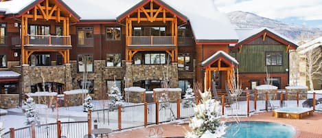Un lodge de varios pisos con zona de piscina, rodeado de árboles nevados y montañas.