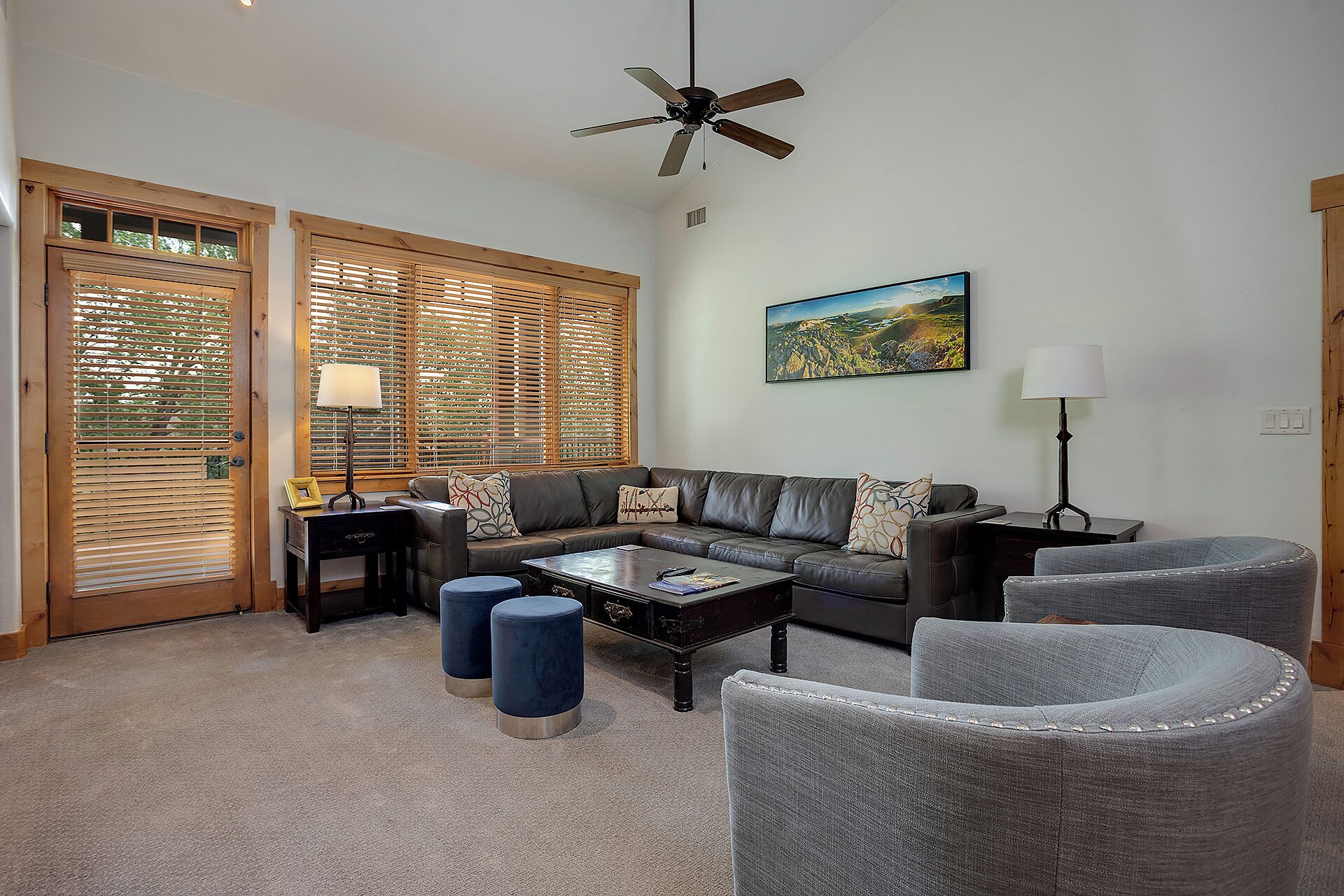 A living room with a sectional sofa, two ottomans, a coffee table, a ceiling fan, and a painting on the wall.
