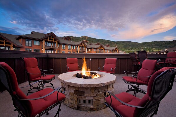 A circular stone fire pit surrounded by red chairs on an outdoor terrace with a view of lodge buildings and hills in the background. - BL6304 | Private Roof Top with Hot Tub | Stunning Condo | Air Chilling | Pools (Steamboat Springs)
