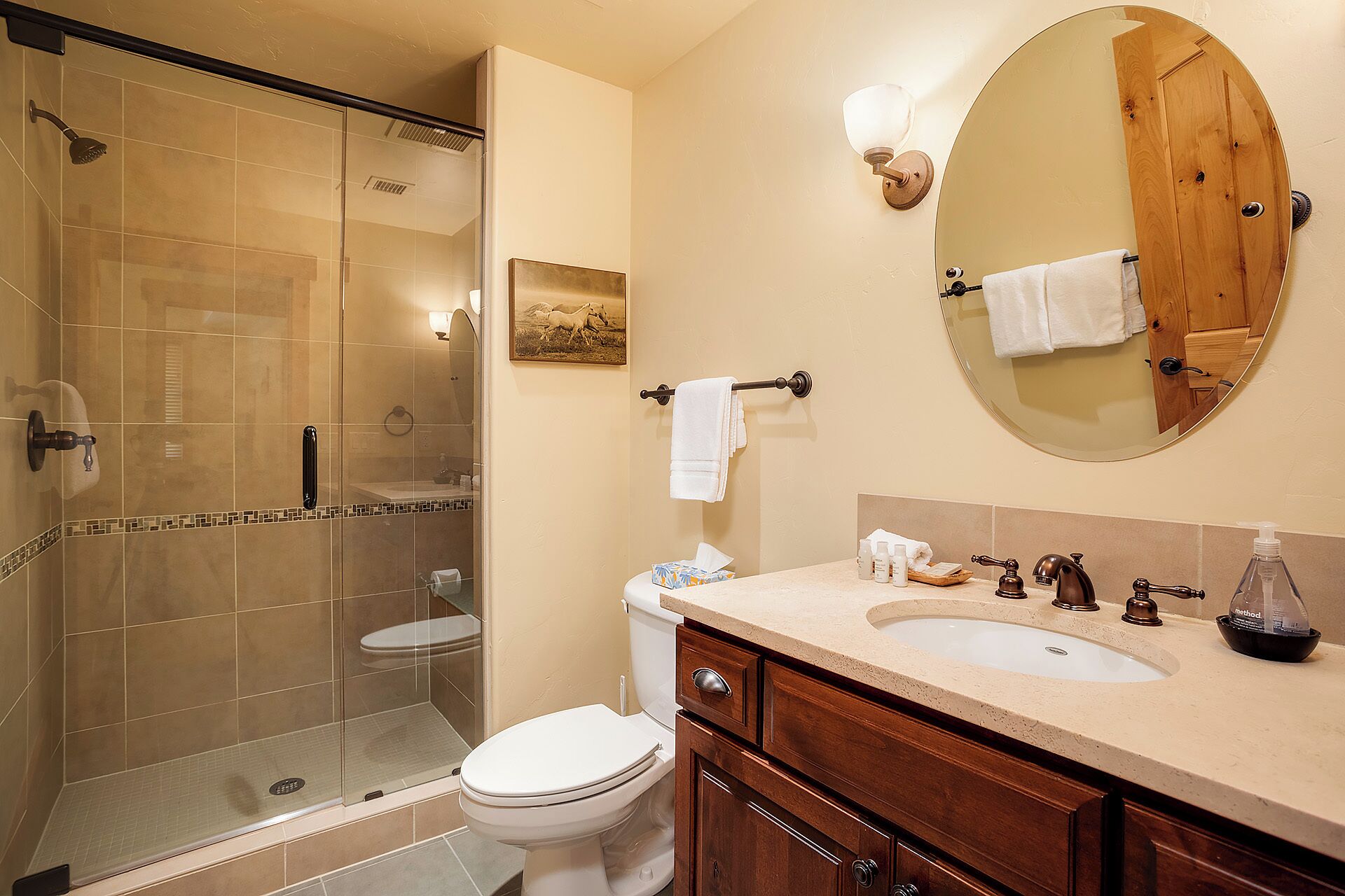 A bathroom with a glass-enclosed shower, a toilet, a sink with toiletries, and a mirror with a wooden door.