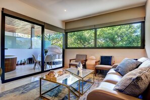 A modern living room with a glass door leading to a balcony, a sofa set, a coffee table, and a view of trees through large windows. - Cruz con Mar PDC (Playa del Carmen)