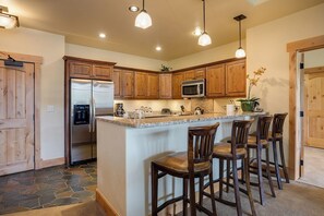 A kitchen with wooden cabinets, a stainless steel refrigerator, a microwave, and a granite countertop. There are three bar stools and a potted plant on the counter. - BL6308 | Top floor Condo with Vaulted Ceilings | Pools | Views | Air Chilling (Steamboat Springs)