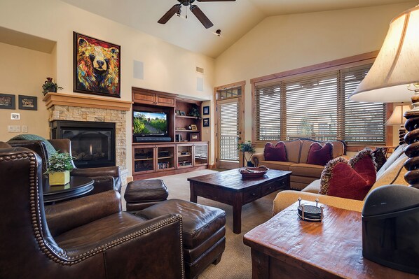 A cozy living room with a fireplace, leather furniture, a television, and a large window with blinds. - BL6308 | Top floor Condo with Vaulted Ceilings | Pools | Views | Air Chilling (Steamboat Springs)