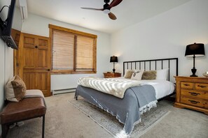 Une chambre à coucher comprenant un lit, des tables de chevet, une armoire en bois, un banc, une fenêtre avec des stores et un ventilateur de plafond.