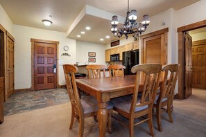 A dining area with a wooden table and chairs, a chandelier, and a kitchen in the background. - CL3105 | Comfortable Mountain Living | A/C | Close to Slopes (Steamboat Springs)