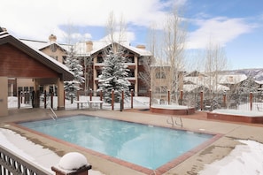 Un espace piscine avec un jacuzzi, entouré d’une terrasse en bois et d’arbres couverts de neige.