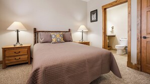 Un dormitorio con una cama, dos mesitas de noche, una mesita auxiliar, una puerta de madera y un baño visible a través de una puerta abierta.