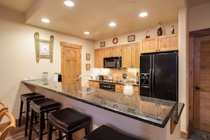 A kitchen with wooden cabinets, a black refrigerator, a microwave, and a granite countertop. There are three stools and a sink. - CL3202 | Cozy Mountain Home | Easy access to Mountain | A/C (Steamboat Springs)