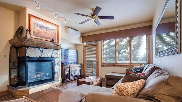 A living room with a stone fireplace, a ceiling fan, and a large window with blinds. - CL3202 | Cozy Mountain Home | Easy access to Mountain | A/C (Steamboat Springs)