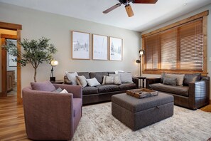 A spacious living room with a brown leather sofa, a brown armchair, and a grey ottoman. There are three framed pictures on the wall, a ceiling fan, and a large window with blinds. - CL3206 | Perfect Condo for a Weekend Getaway | A/C | Close to Slopes (Steamboat Springs)