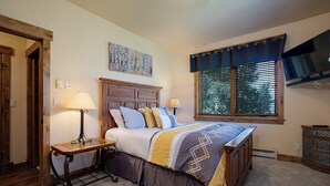 Un dormitorio con cama de madera, dos lámparas de noche, una ventana con persianas y un televisor en la pared.