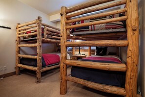 Twee stapelbedden met houten frames en meerdere kussens, in een kamer met vloerbedekking en een kleine tafel.