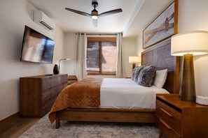 Ein Schlafzimmer mit einem Bett, Nachttischen, einem Schreibtisch, einem Deckenventilator, einem Fernseher und einem Fenster mit Jalousien.