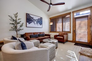 Ein geräumiges Wohnzimmer mit einem braunen Ledersofa, einem beigefarbenen Sessel und einem Couchtisch. Es gibt ein großes Fenster mit Jalousien, einen Deckenventilator und ein gerahmtes Kunstwerk an der Wand.