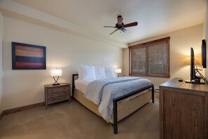 Un dormitorio con cama, mesitas de noche, ventilador de techo, ventana con persianas y un cuadro en la pared.