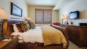 Quarto com cama, cabeceira de madeira, mesas de cabeceira, cômoda, televisão e janela com persianas.