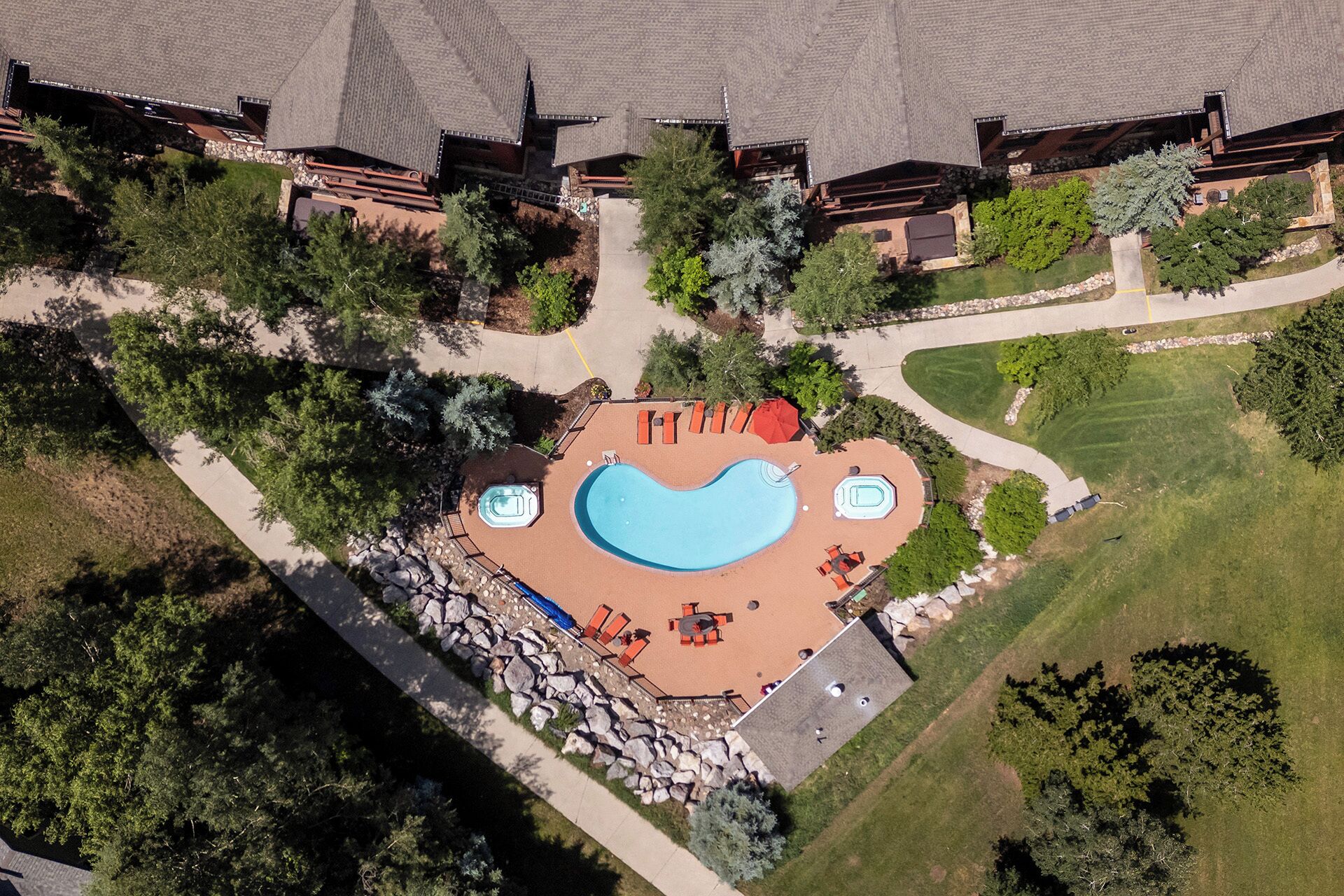 An aerial view of a resort with a large swimming pool, a hot tub, and outdoor seating areas surrounded by trees and a well-maintained lawn.
